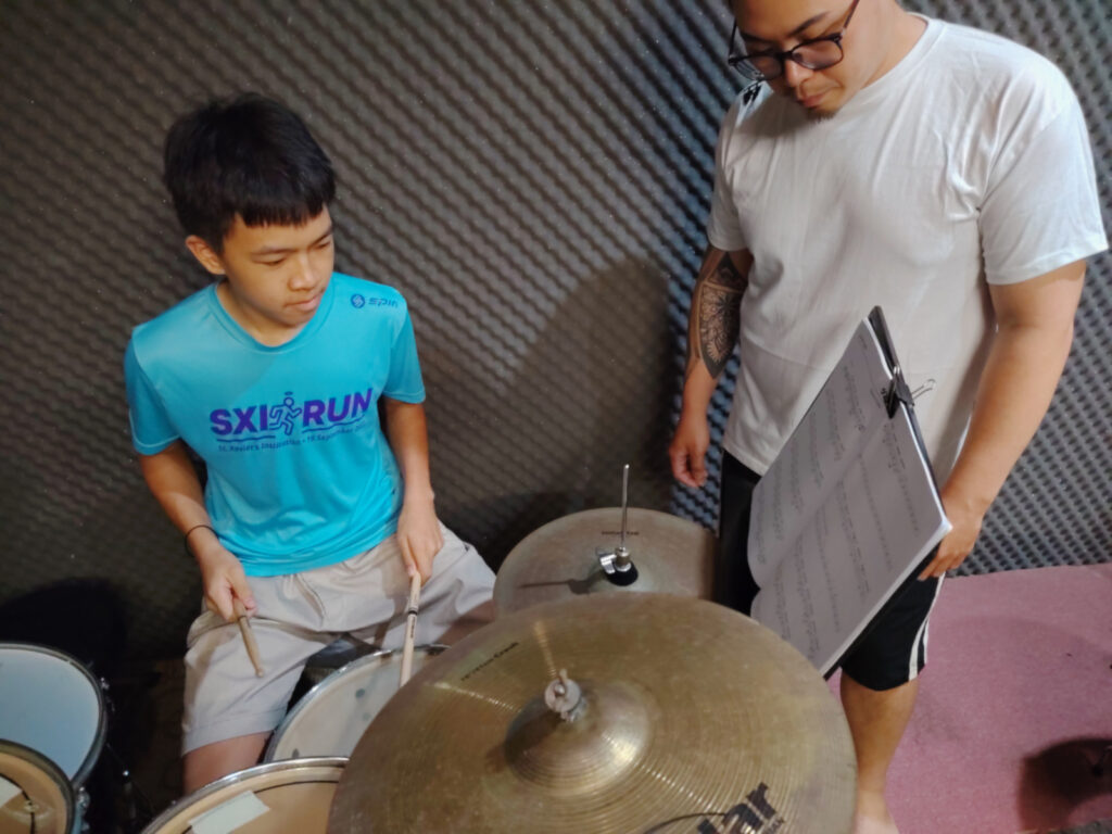 A student plays the drums while reading a music sheet, with a teacher standing beside them and guiding their performance.