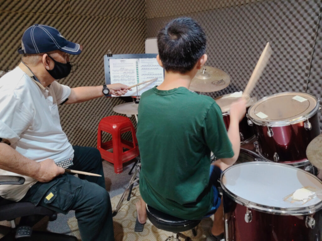Teacher sitting beside student during drum lesson, pointing at sheet music